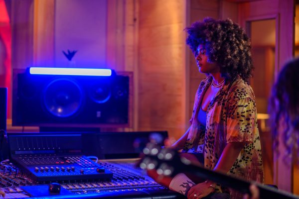A musician standing next to a mixing console in a studio with a guitar player